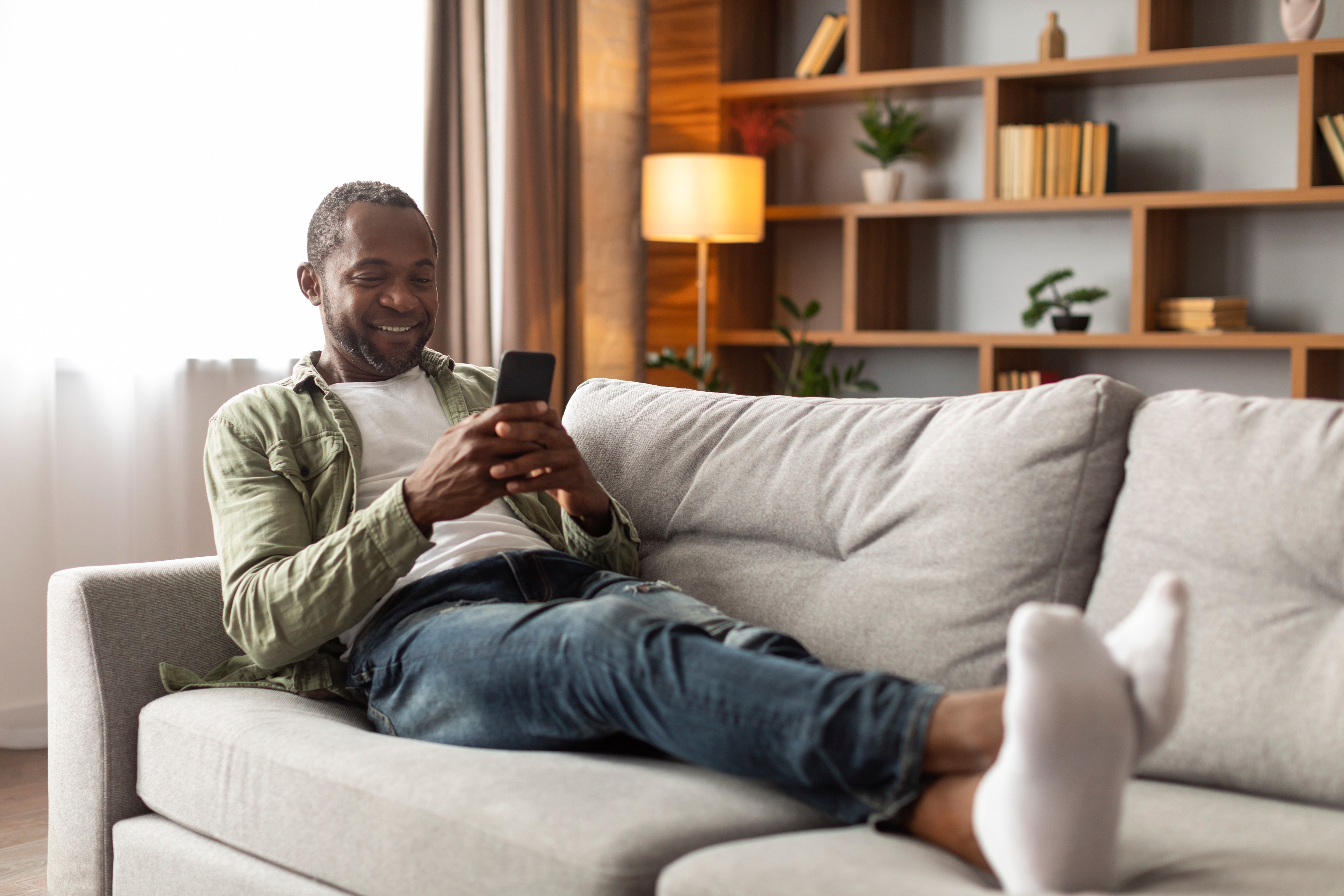 Picture of a man sitting on a couch with his legs up staring at his phone with joy.